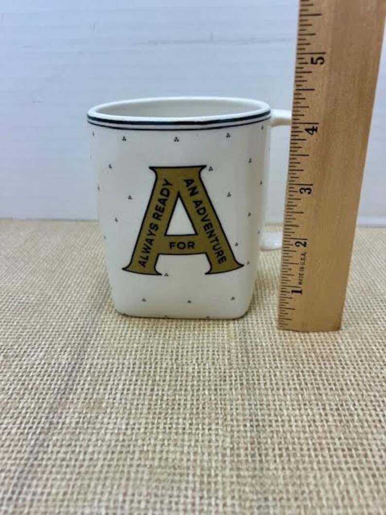 Puede incluir: Taza de cer&aacute;mica blanca con ribete negro y la letra "A" dorada, con las palabras "ALWAYS READY FOR AN ADVENTURE". La taza tiene peque&ntilde;os puntos negros y una regla de madera al lado.