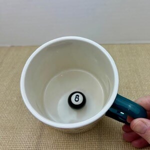 May include: A white ceramic mug with a teal handle, featuring a black and white eight-ball at the bottom. The mug is set on a textured, tan surface.