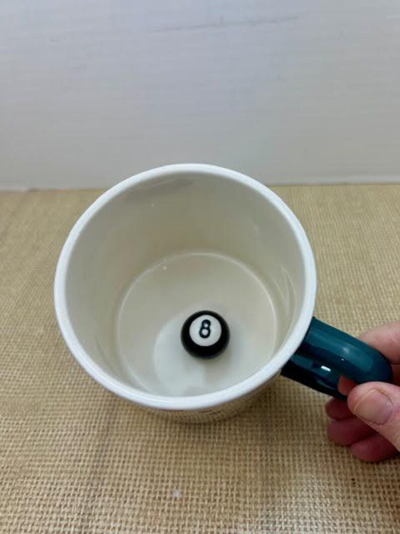 May include: A white ceramic mug with a teal handle, featuring a black and white eight-ball at the bottom. The mug is set on a textured, tan surface.