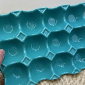 May include: A turquoise ceramic egg holder with twelve individual compartments. The holder has a scalloped edge and geometric details, including square and triangular accents. The glossy finish reflects light, highlighting the aqua color.