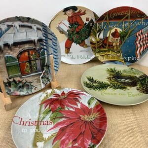 May include: Set of six decorative ceramic plates featuring various Christmas themes. The plates feature images of a snow-covered house, a poinsettia flower, a bird in a nest, a Santa Claus figure, and a child holding a gift. The plates have a white background with colorful illustrations and text.