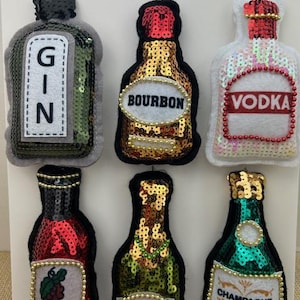 May include: Six sequined bottle ornaments: Gin, Bourbon, Vodka, Red Wine, White Wine, and Champagne. Each ornament is shaped like a bottle with a label and decorative accents. The ornaments are attached to a white display board.