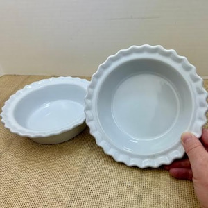 May include: Two white ceramic pie dishes with scalloped edges. The dishes are round with a shallow depth and a glossy finish. One dish is in the foreground, and the other is slightly behind it.