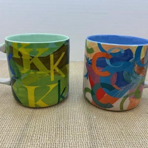 May include: Two ceramic mugs. One is green with yellow "K" letters. The other is white with colorful abstract shapes. Both mugs have white handles and rims. The mugs are on a tan surface.