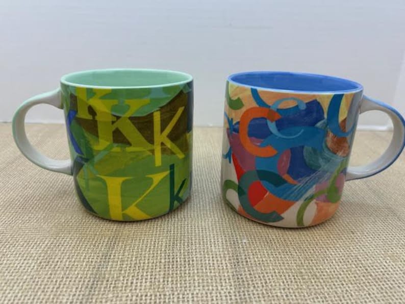 May include: Two ceramic mugs. One is green with yellow "K" letters. The other is white with colorful abstract shapes. Both mugs have white handles and rims. The mugs are on a tan surface.