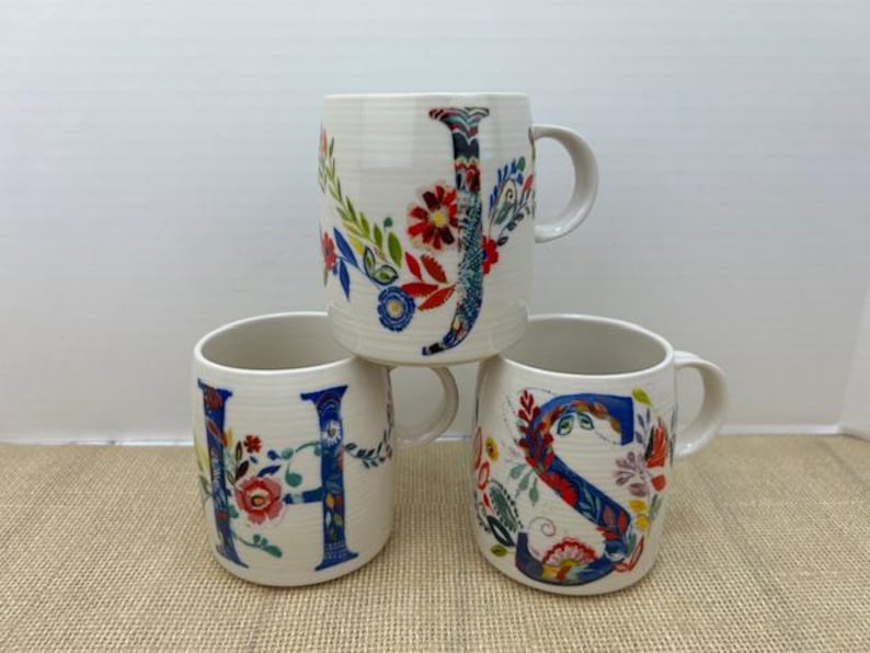 Puede incluir: Tres tazas de cer&aacute;mica blanca con dise&ntilde;os florales e iniciales. Las tazas presentan las letras "J", "H" y "S" en azul, con coloridos estampados florales. Cada taza tiene un asa y una superficie texturizada.