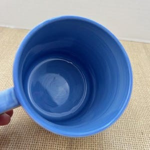 May include: A light blue ceramic mug with a handle. The interior and exterior of the mug are a solid light blue color. The mug is empty and appears to be clean.