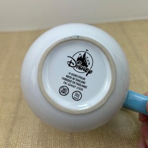 Op de afbeelding: Een witte keramische mok met een blauwe handgreep. De onderkant van de mok heeft een zwart Disney-logo met de tekst "Disney/Pixar Gemaakt in Thailand Fabrique en Thailande FAC-024367-21020".