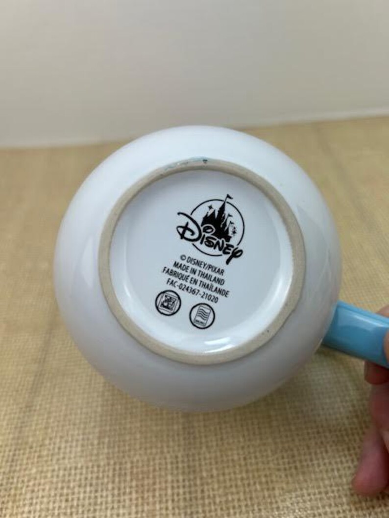 Op de afbeelding: Een witte keramische mok met een blauwe handgreep. De onderkant van de mok heeft een zwart Disney-logo met de tekst "Disney/Pixar Gemaakt in Thailand Fabrique en Thailande FAC-024367-21020".