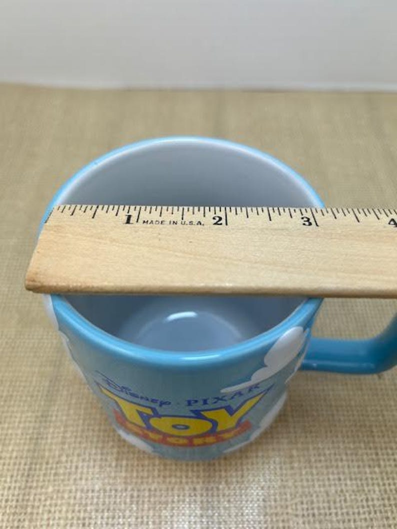 Op de afbeelding: Een blauwe keramische mok met een wit wolkenontwerp en de woorden "Disney Pixar Toy Story" erop gedrukt. De mok heeft een diameter van 7,6 cm.