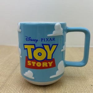 Op de afbeelding: Een blauwe keramische mok met een witte handgreep. De mok heeft een wit wolkenontwerp en de woorden "Disney Pixar Toy Story" in blauw, geel en rood.
