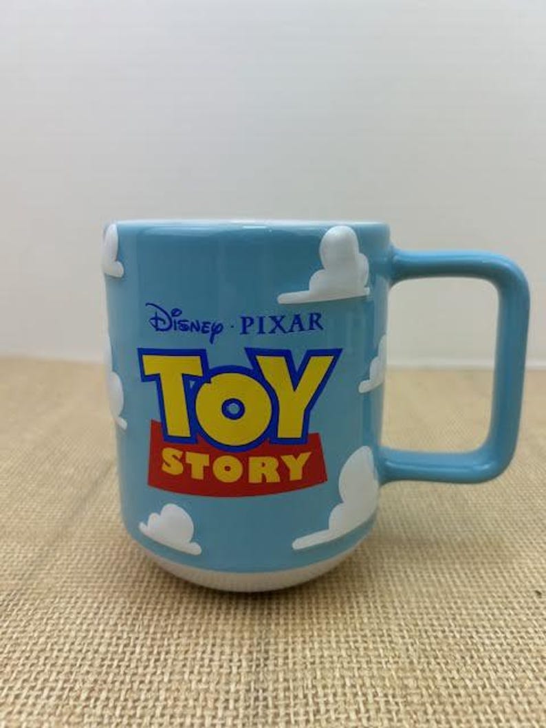 Op de afbeelding: Een blauwe keramische mok met een witte handgreep. De mok heeft een wit wolkenontwerp en de woorden "Disney Pixar Toy Story" in blauw, geel en rood.