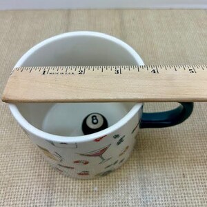 May include: White ceramic mug with a dark green handle, featuring a black eight ball inside. The mug is decorated with colorful cocktail and food illustrations. A wooden ruler is placed across the top of the mug.