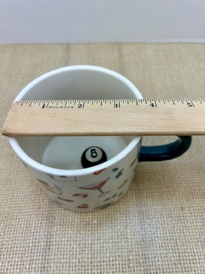 May include: White ceramic mug with a dark green handle, featuring a black eight ball inside. The mug is decorated with colorful cocktail and food illustrations. A wooden ruler is placed across the top of the mug.