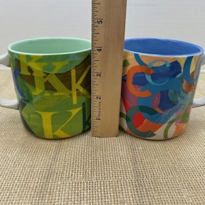 May include: Two ceramic mugs with handles. One mug is green with yellow and brown abstract designs. The other mug has a blue rim and is decorated with colorful abstract shapes. A wooden ruler is placed between the mugs.