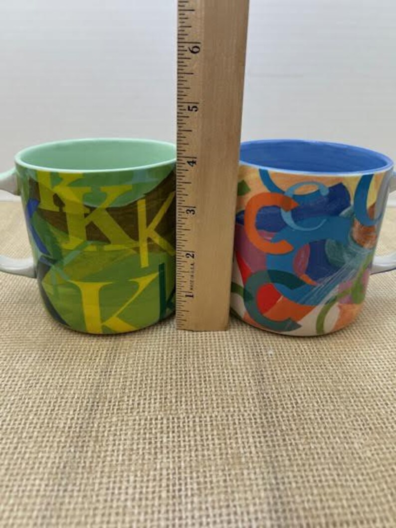 May include: Two ceramic mugs with handles. One mug is green with yellow and brown abstract designs. The other mug has a blue rim and is decorated with colorful abstract shapes. A wooden ruler is placed between the mugs.