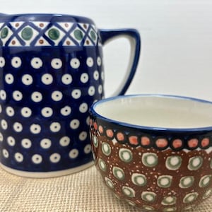 May include: A blue and white ceramic pitcher and cup set. The pitcher has a handle and spout, decorated with white dots and geometric patterns. The cup has a handle and a brown and white pattern with green and orange accents.
