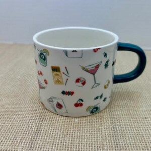 May include: White ceramic mug with a dark teal handle, decorated with colorful illustrations of cocktails, playing cards, cherries, and other game-themed designs. The mug is sitting on a textured, tan surface.