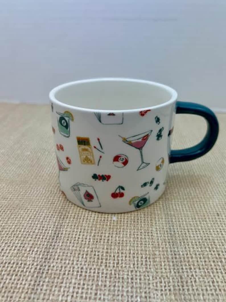 May include: White ceramic mug with a dark teal handle, decorated with colorful illustrations of cocktails, playing cards, cherries, and other game-themed designs. The mug is sitting on a textured, tan surface.
