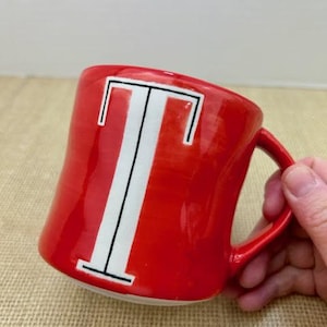 May include: A red ceramic mug with a white letter "T" in the center. The letter is outlined in black. The mug has a red handle and a glossy finish. The mug is on a tan surface.