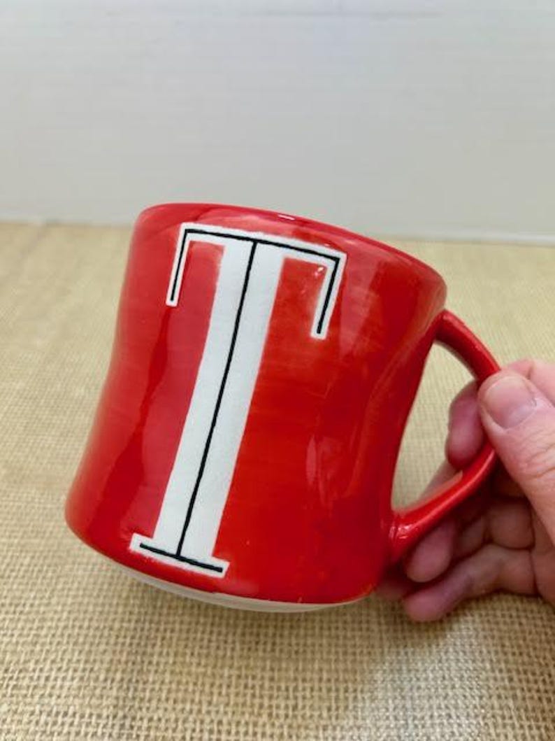 May include: A red ceramic mug with a white letter "T" in the center. The letter is outlined in black. The mug has a red handle and a glossy finish. The mug is on a tan surface.