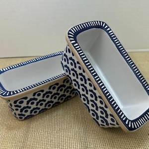 May include: Two rectangular ceramic loaf pans. The pans have a white interior and a beige exterior with a navy blue geometric pattern. The rims are navy blue with white accents. The pans are on a textured, tan surface.