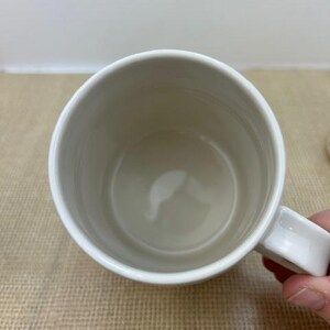 Puede incluir: Una taza de cer&aacute;mica blanca con asa, vista desde arriba. El interior de la taza es de color beige claro. La taza est&aacute; sujeta por una mano.