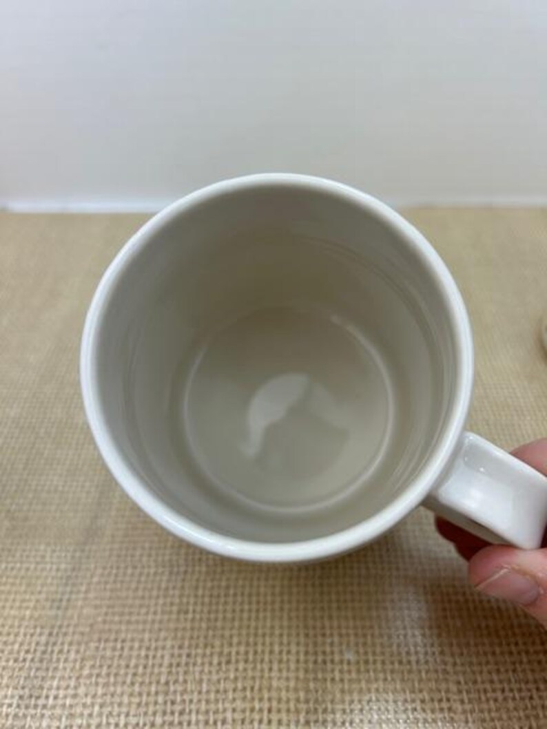 Puede incluir: Una taza de cer&aacute;mica blanca con asa, vista desde arriba. El interior de la taza es de color beige claro. La taza est&aacute; sujeta por una mano.