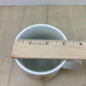 May include: White ceramic mug with a handle, measured with a wooden ruler. The ruler reads "MADE IN U.S.A." and measures approximately 7.6 cm across the top of the mug. The mug sits on a tan surface.