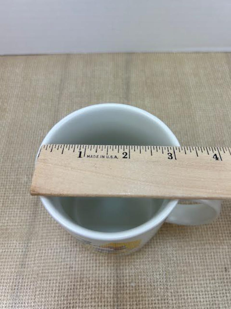 May include: White ceramic mug with a handle, measured with a wooden ruler. The ruler reads "MADE IN U.S.A." and measures approximately 7.6 cm across the top of the mug. The mug sits on a tan surface.