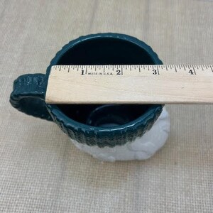 Pu&ograve; includere: Una tazza in ceramica verde scuro con base bianca, di circa 10 cm di diametro. La tazza presenta un design strutturato, simile a un tessuto a maglia, attorno alla parte superiore e al manico. Un righello di legno &egrave; appoggiato sull'apertura della tazza.