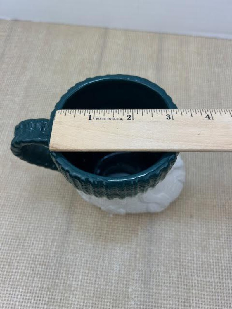 Pu&ograve; includere: Una tazza in ceramica verde scuro con base bianca, di circa 10 cm di diametro. La tazza presenta un design strutturato, simile a un tessuto a maglia, attorno alla parte superiore e al manico. Un righello di legno &egrave; appoggiato sull'apertura della tazza.