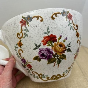 May include: A large white ceramic teacup with a floral design. The cup has a speckled glaze and a handle. The floral design features a variety of flowers in shades of red, yellow, purple, and green.