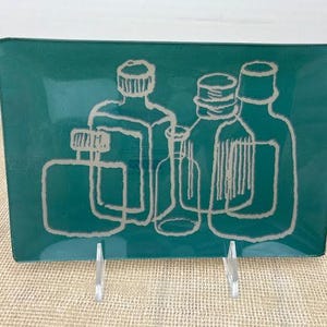May include: Teal rectangular decorative tray with a white outline design of various bottles. The tray is supported by two clear plastic stands, and sits on a tan surface.