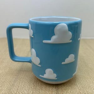 Op de afbeelding: Een lichtblauwe keramische mok met een witte handgreep. De mok heeft een herhalend patroon van witte wolken op een lichtblauwe achtergrond.