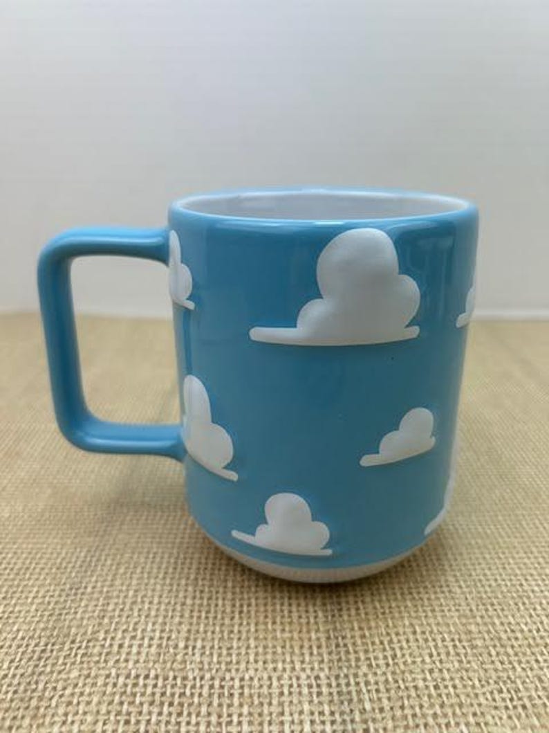 Op de afbeelding: Een lichtblauwe keramische mok met een witte handgreep. De mok heeft een herhalend patroon van witte wolken op een lichtblauwe achtergrond.