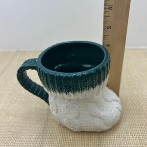 Pu&ograve; includere: Una tazza in ceramica a forma di calzino, con interno e manico verde scuro. Il bordo superiore &egrave; verde, che passa a un design a trecce bianco. Un righello di legno &egrave; accanto alla tazza.