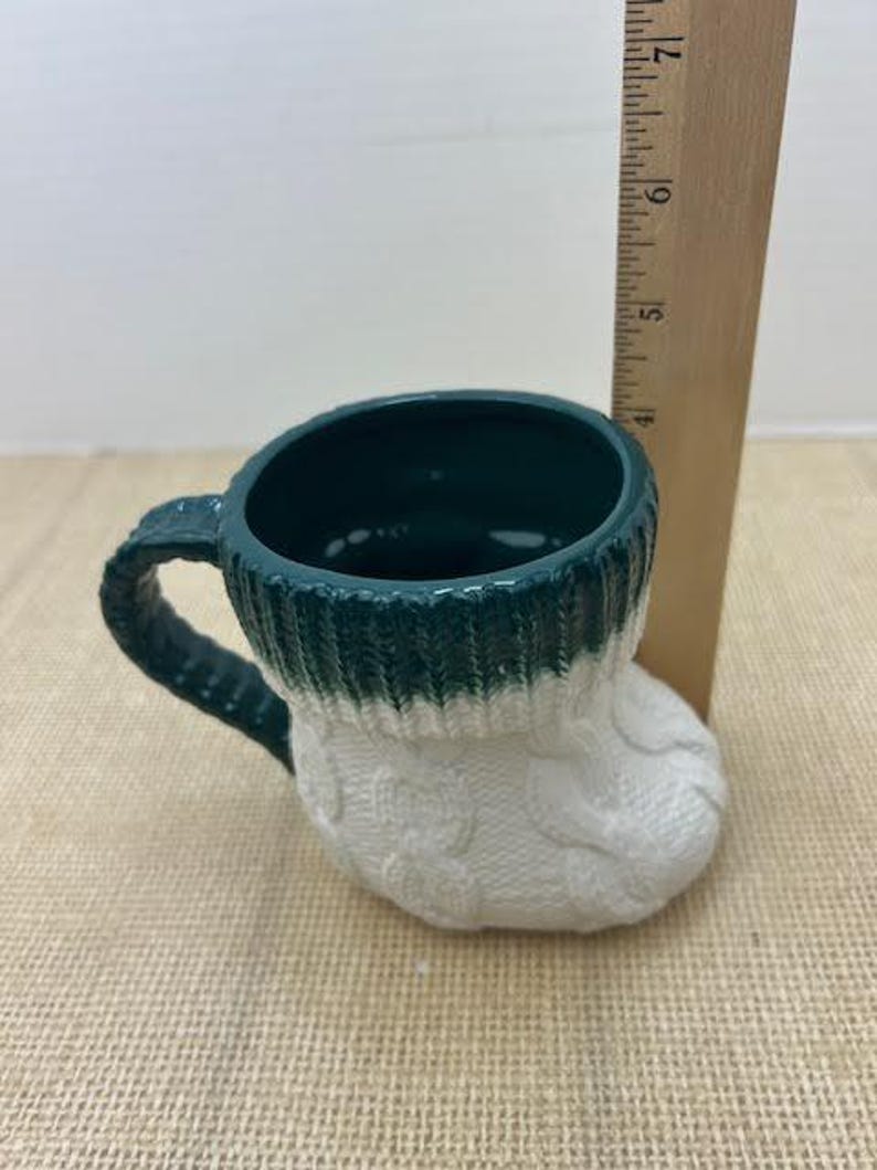 Pu&ograve; includere: Una tazza in ceramica a forma di calzino, con interno e manico verde scuro. Il bordo superiore &egrave; verde, che passa a un design a trecce bianco. Un righello di legno &egrave; accanto alla tazza.