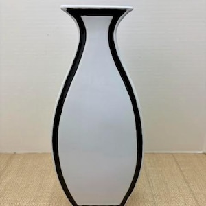 May include: A tall, white ceramic vase with a smooth, glossy finish. The vase features a distinct, bold black outline that follows the curved silhouette of the body and flares slightly at the top rim, creating a clean, high-contrast graphic design.