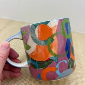 May include: A ceramic mug with a white handle and a colorful abstract design. The design features overlapping shapes in orange, green, purple, and blue. The mug is held by a hand.
