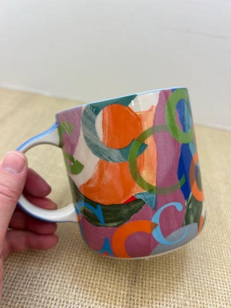 May include: A ceramic mug with a white handle and a colorful abstract design. The design features overlapping shapes in orange, green, purple, and blue. The mug is held by a hand.