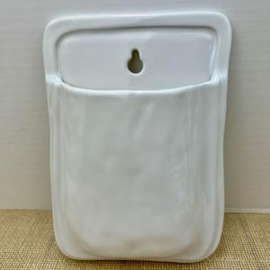 May include: A white ceramic wall pocket with a glossy finish and a textured, organic surface. The rectangular piece features a rounded bottom and a single keyhole opening at the top center for mounting, designed to hold small items or plants.