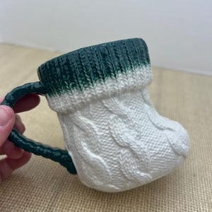 Pu&ograve; includere: Una tazza in ceramica a forma di calzino, con un motivo a trecce bianco e un bordo e manico verde scuro. La tazza &egrave; progettata per assomigliare a un caldo calzino invernale.