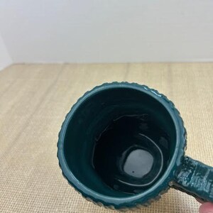 Pu&ograve; includere: Una tazza in ceramica verde acqua con esterno strutturato e manico. La tazza ha un interno scuro ed &egrave; appoggiata su una superficie beige. La tazza &egrave; vuota.