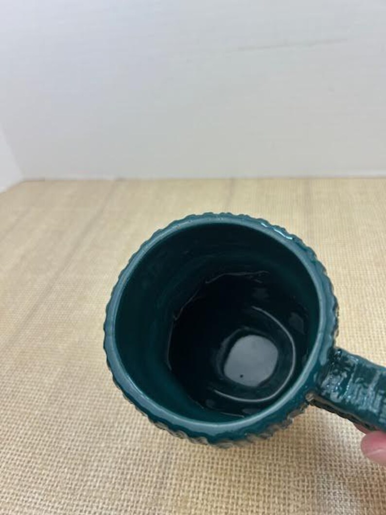 Pu&ograve; includere: Una tazza in ceramica verde acqua con esterno strutturato e manico. La tazza ha un interno scuro ed &egrave; appoggiata su una superficie beige. La tazza &egrave; vuota.