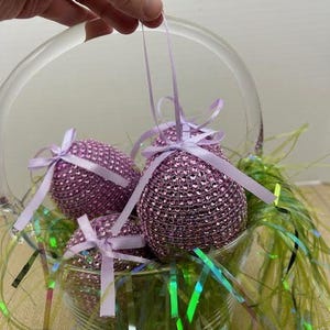 May include: A glass basket filled with four lavender-colored Easter eggs decorated with rhinestones. The eggs are tied with lavender ribbons and are sitting on a bed of green and iridescent green grass.