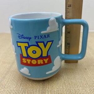Op de afbeelding: Een lichtblauwe keramische mok met een wit wolkenontwerp. De mok heeft het Disney Pixar Toy Story logo in geel, rood en blauw.