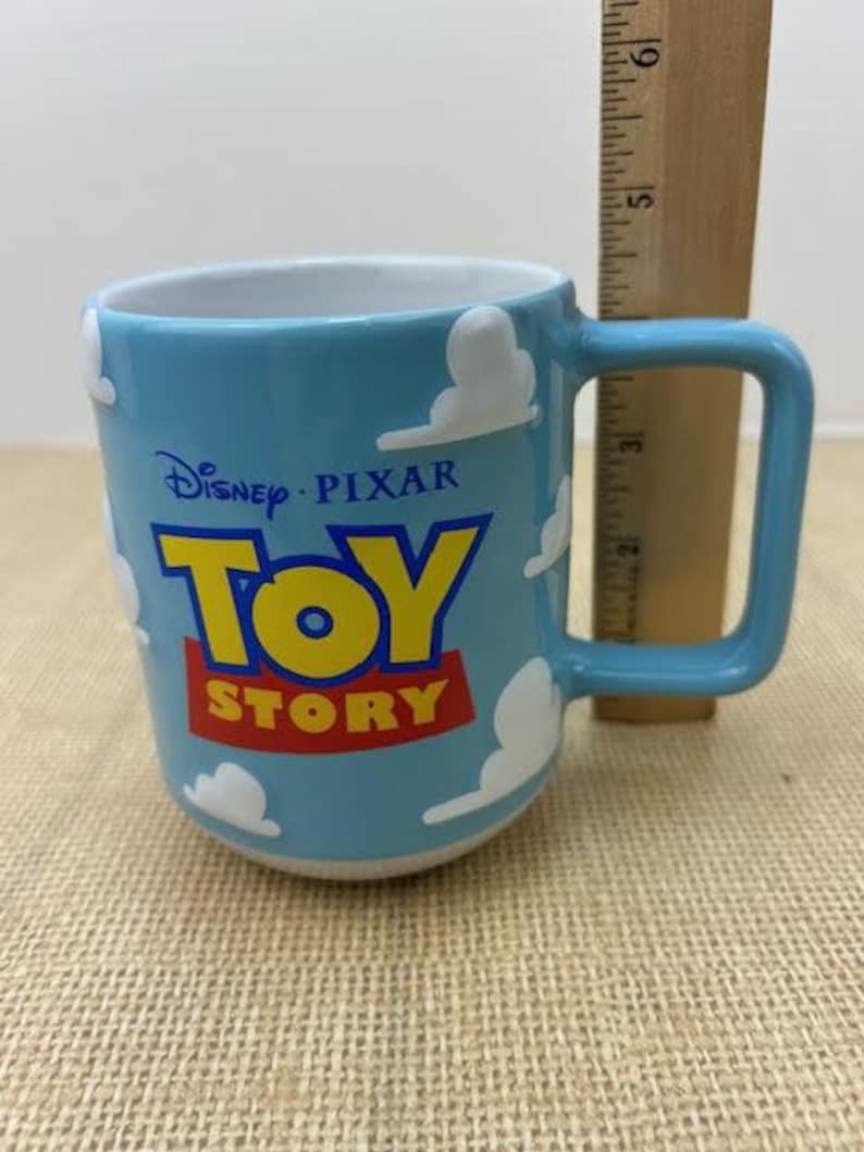 Op de afbeelding: Een lichtblauwe keramische mok met een wit wolkenontwerp. De mok heeft het Disney Pixar Toy Story logo in geel, rood en blauw.