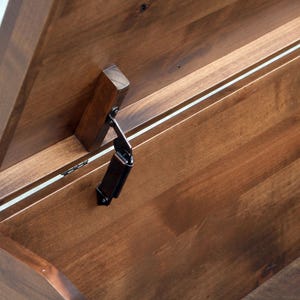 May include: Close-up of a wooden chest with the lid partially open. The chest is made of stained wood with visible grain and a dark finish. A metal hinge and wooden support are visible.