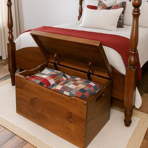 May include: A wooden storage chest with an open lid, revealing two patchwork cushions. The chest is placed at the foot of a wooden bed with a red blanket and white pillows. The room has a neutral color palette.
